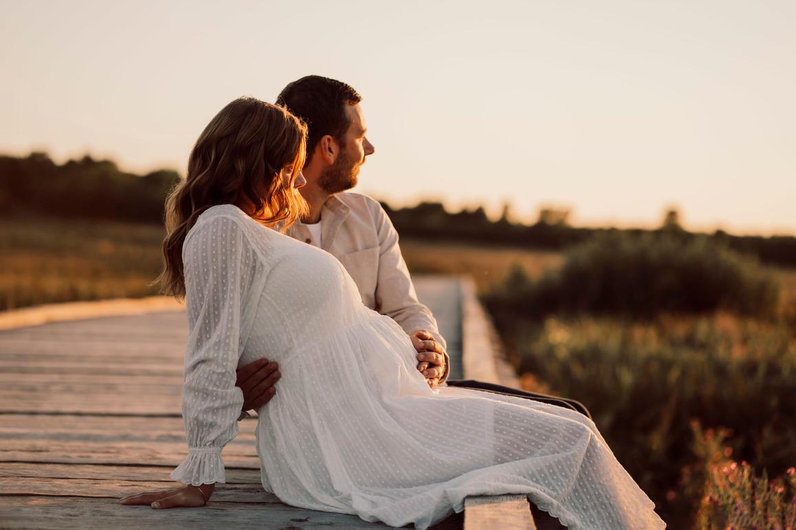Et par sidder på en træbro ved solnedgang, kvinden er gravid og bærer en hvid kjole.