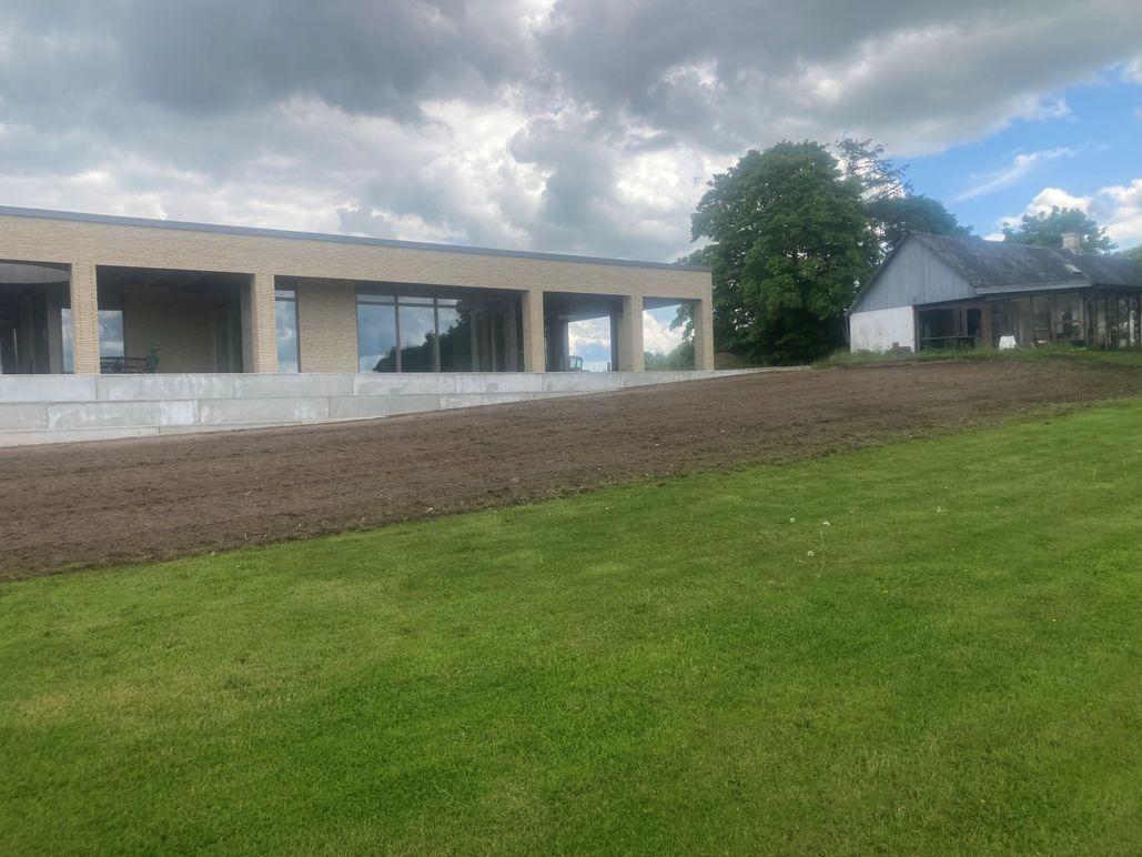 Moderne hus med store vinduer og græsplæne, omgivet af træer og en skyet himmel.