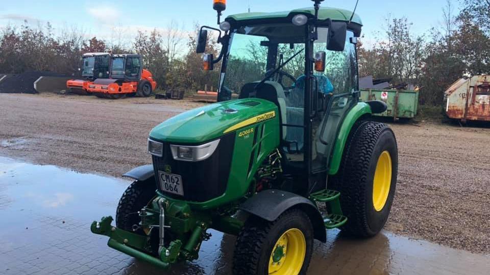 En grøn traktor parkeret på en gård med grusbelægning og andre landbrugsmaskiner i baggrunden.