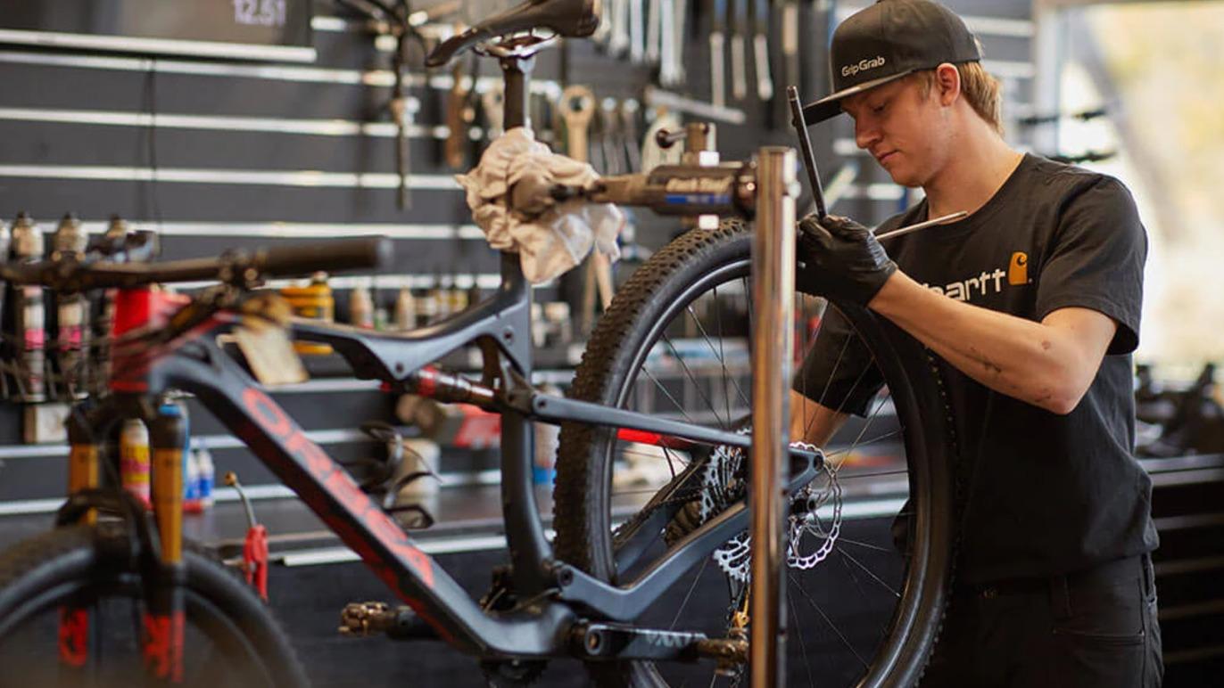 En mekaniker arbejder på en cykel i en cykelbutik med værktøj og udstyr i baggrunden.