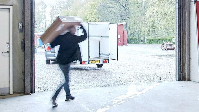 En person bærer en kasse og går hurtigt ud af en garage, mens en trailer står åben i baggrunden.