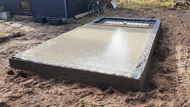 Ny betonplade, der er blevet hældt og glattet, ligger på en sandet grund ved en bygning.
