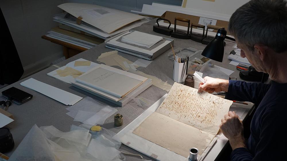 En person arbejder med papir og kunstmaterialer på et bord fyldt med skitser og værktøjer.