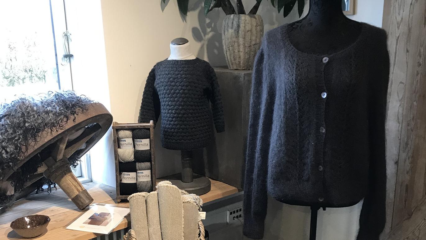 En striktrøje og en strikbluse hænger på en mannequin, omgivet af garn og strikkeudstyr i en hyggelig indretning.
