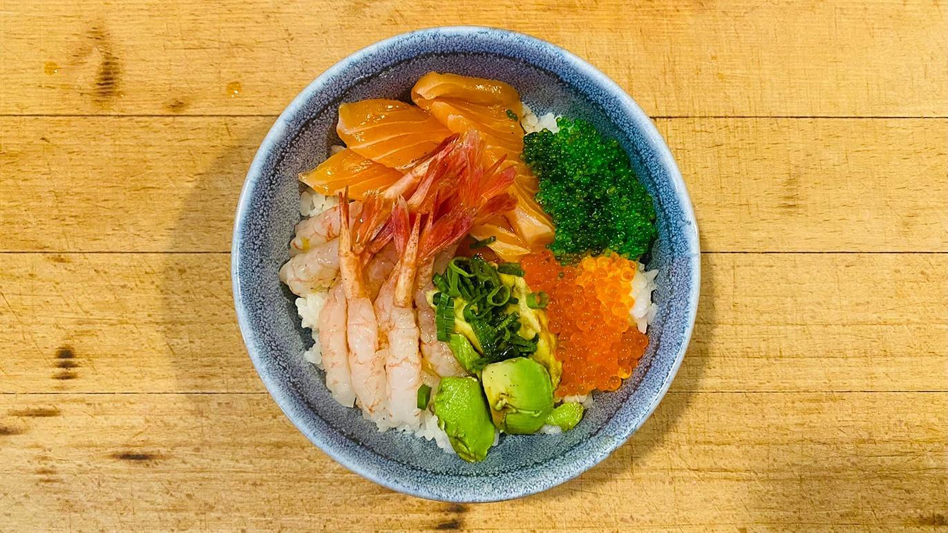 En skål med sushi bestående af ris, laks, rejer, avocado, tang og fiskeæg på et træbord.