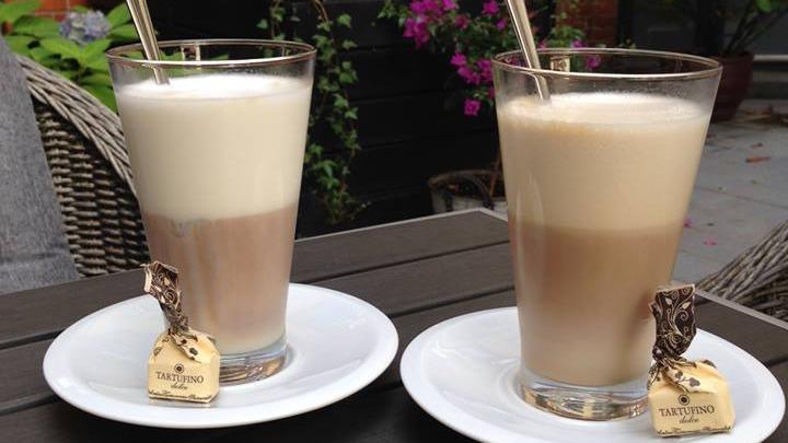 To kopper med latte, serveret i glas med skum og chokolade, placeret på hvide tallerkener.