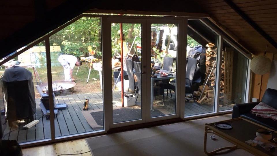 Udsigt fra stuen med store vinduer, der viser en have med møbler og en person, der arbejder.