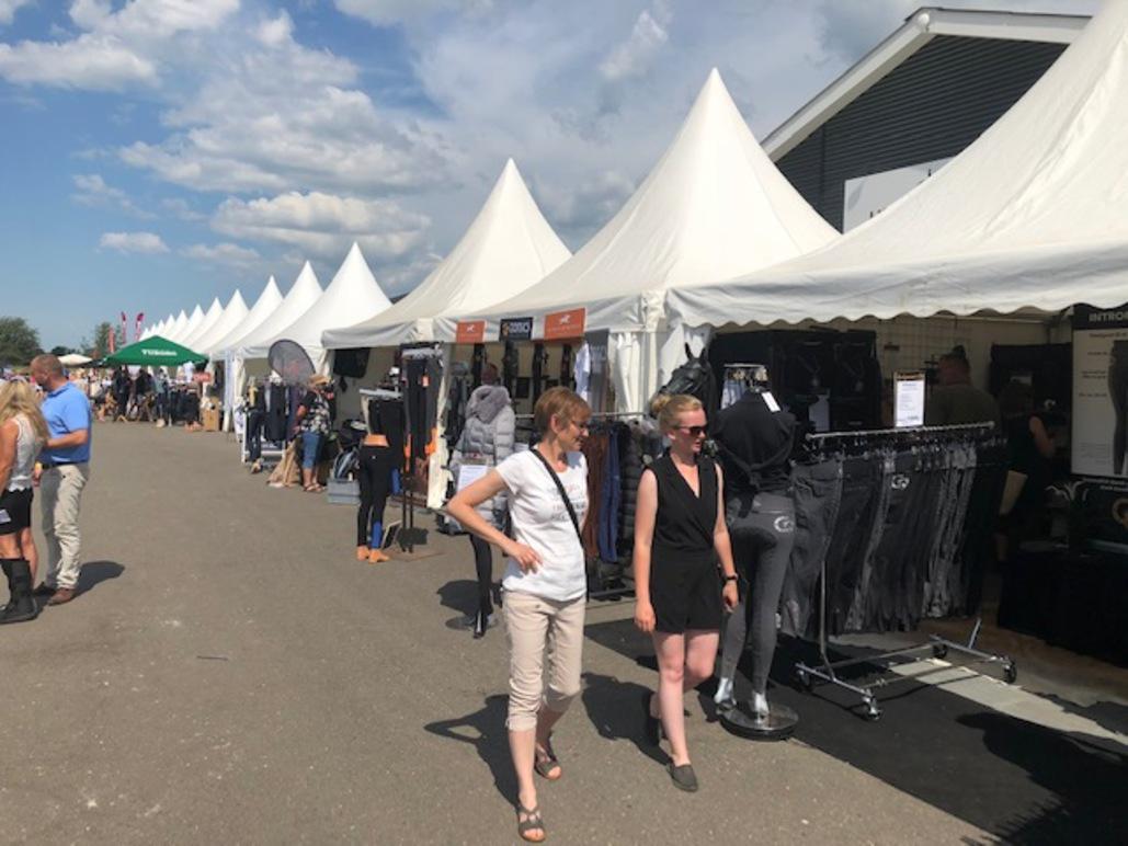 Mennesker går forbi boder med telte på en udendørs messe under en blå himmel med skyer.