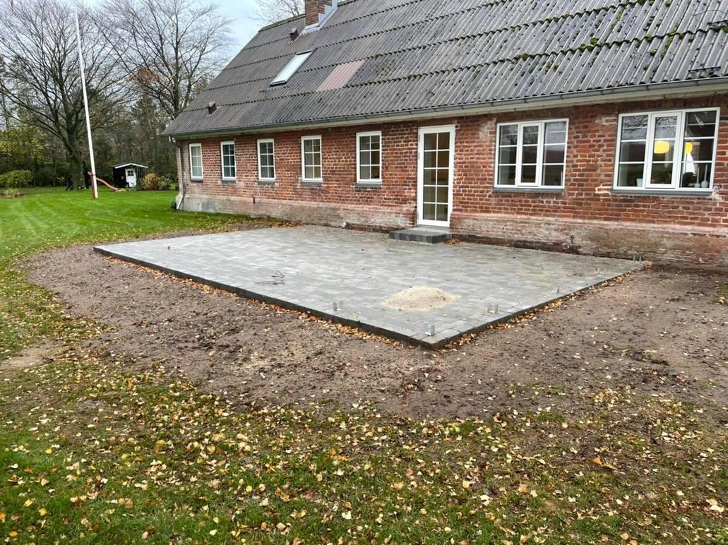 Hus med ny flisebelægning i haven, omgivet af græs og træer, med en grusomgivet terrasse.