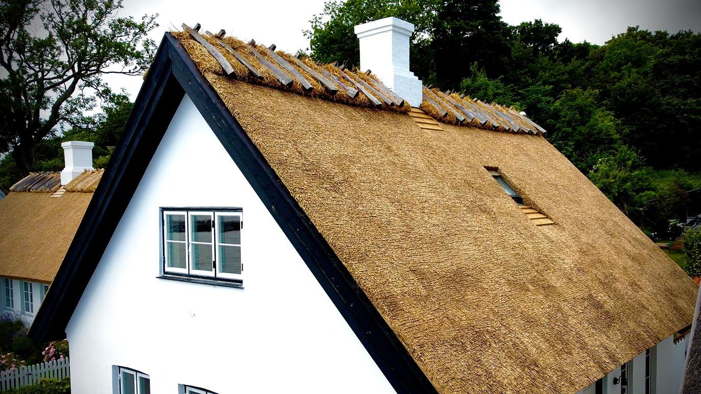 Et hvidt hus med stråtag og vinduer, omgivet af grønne træer og en klar himmel.