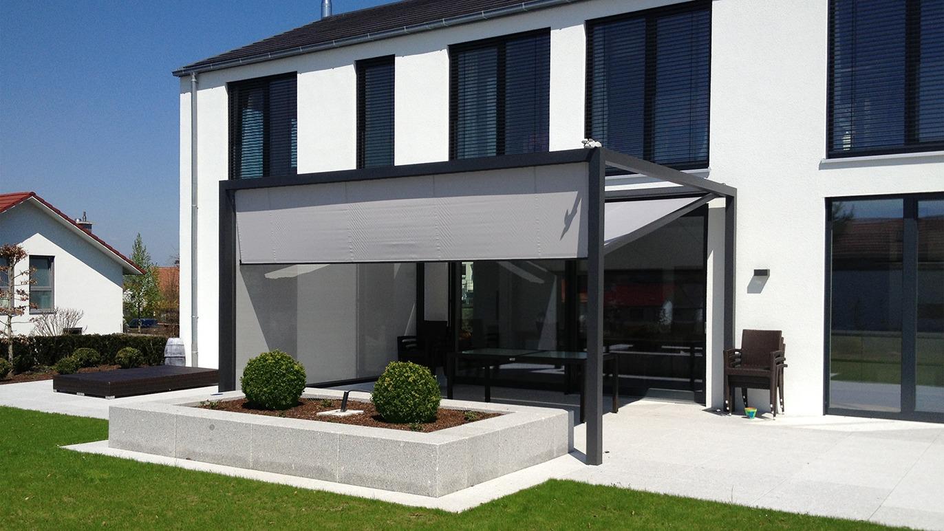 Moderne hus med terrasseoverdækning og grøn græsplæne, omgivet af buske og en stenbed.