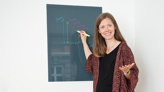 Kvinde skriver med farvet kridt på en tavle og smiler, mens hun gestikulerer med den anden hånd.