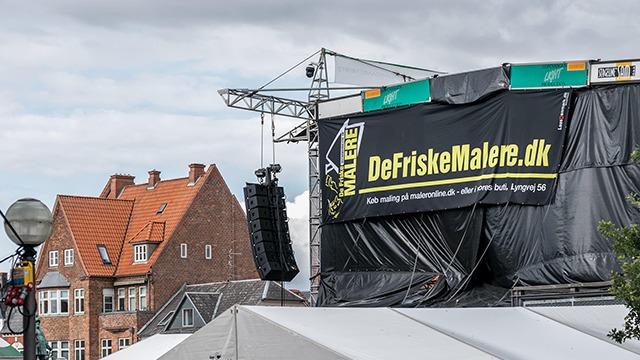 En scene med et banner fra DeFriskeMalere.dk og bygninger i baggrunden under en overskyet himmel.