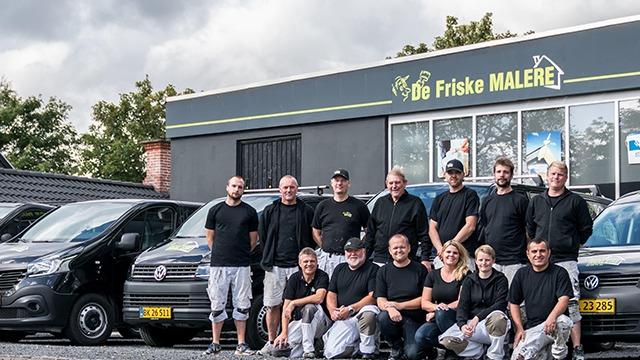 En gruppe malere står foran deres firma, De Friske Malere, med biler i baggrunden.