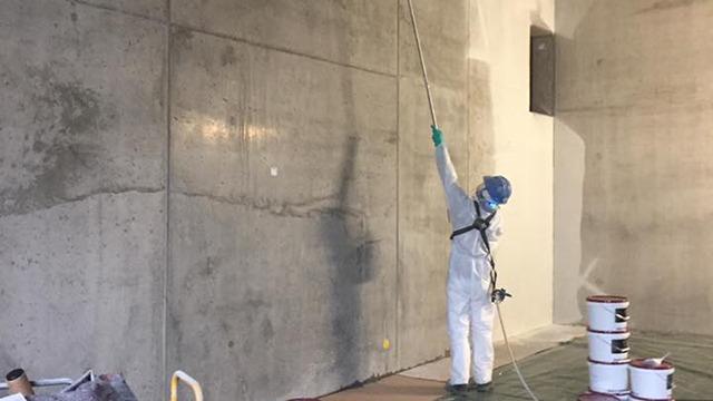 En maler i beskyttelsesdragt sprøjter maling på en betonvæg i et stort rum med malerbøtter omkring.