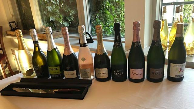 En række champagneflasker i forskellige farver og størrelser, præsenteret på et bord ved et vindue.