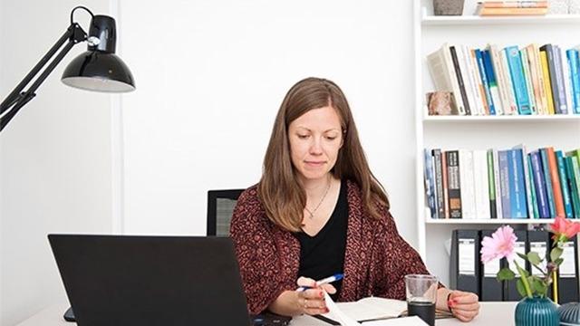 Kvinde sidder ved skrivebord med bærbar computer, skriver i notesbog og har blomster og bøger i baggrunden.