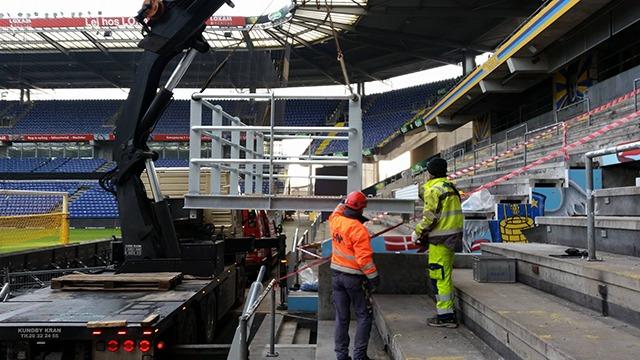 Arbejdsfolk flytter udstyr ind på en stadiontribune med en kran og lastbil i baggrunden.