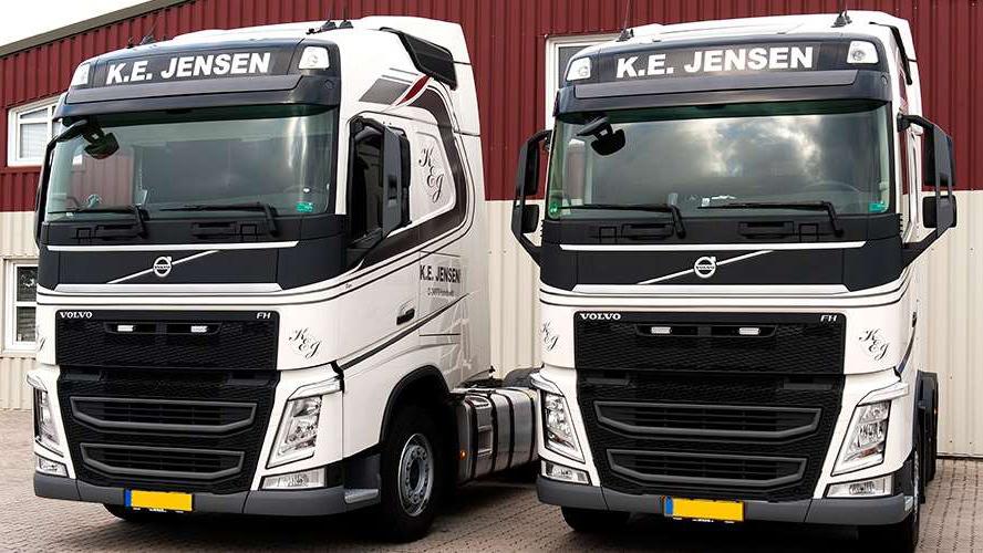 To lastbiler fra K.E. Jensen parkeret foran en bygning, begge i hvid og sort farve med Volvo-logo.