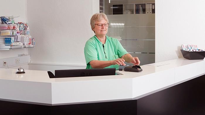En receptionist i en grøn skjorte står ved en moderne skranke med en computer og kontorartikler i baggrunden.