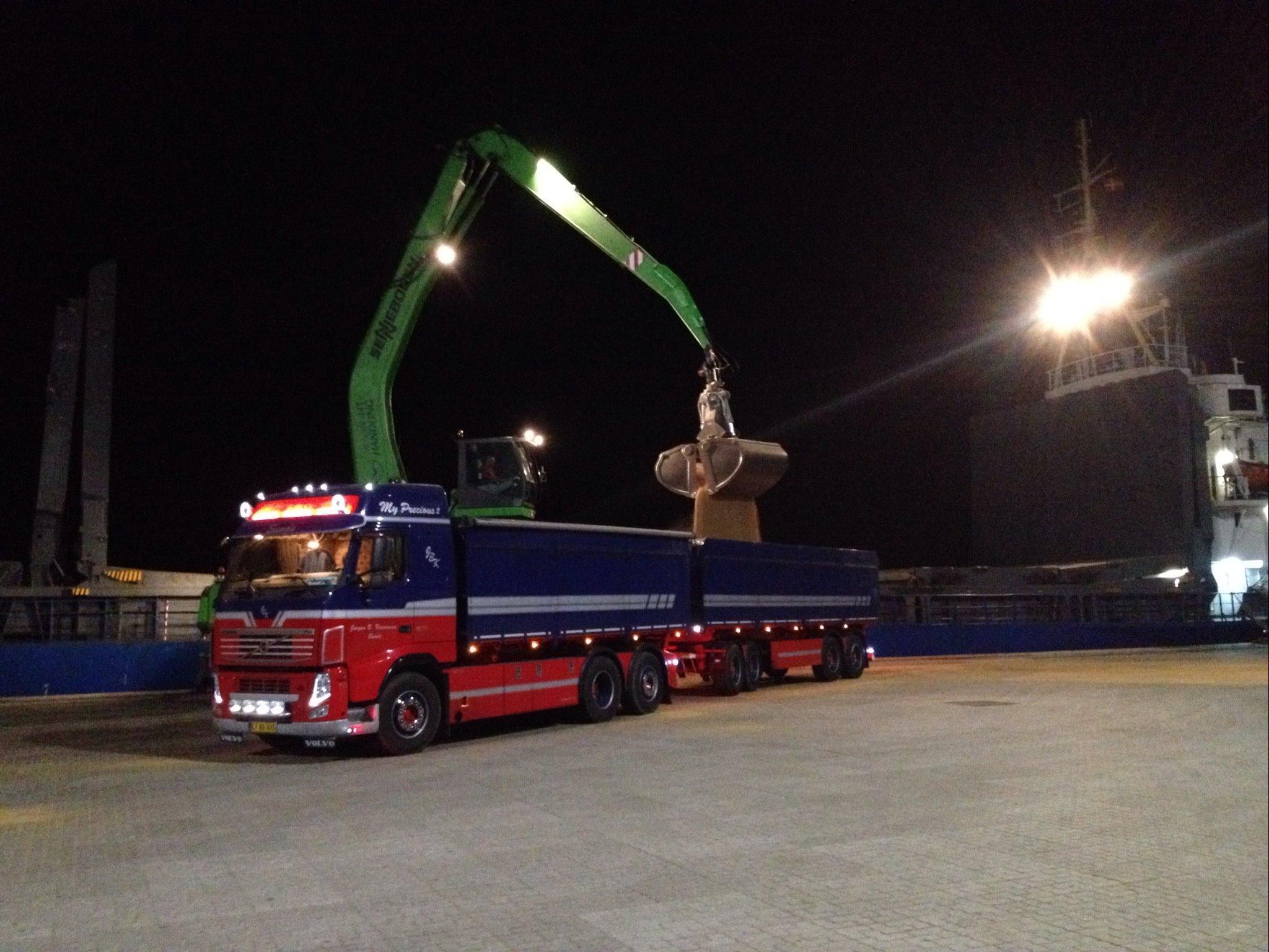 Lastbil med kran, der løfter en stor metalgenstand på havnen om natten.