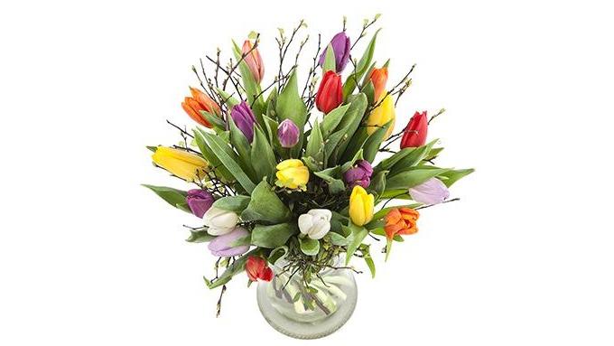 En farverig buket tulipaner i en glasvase med grønne blade og grene.