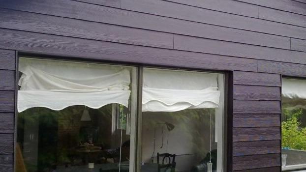 Facade af et hus med mørkt træpanel og hvide markiser ved vinduerne.