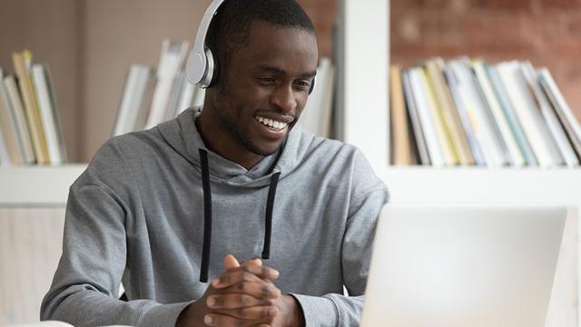En mand med hovedtelefoner smiler, mens han ser på sin bærbare computer i et lyst rum med bøger i baggrunden.