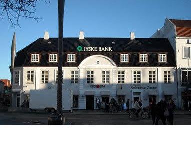 Jyske Bank bygning med hvid facade og sort tag, beliggende på en travl gade med cyklister og forbipasserende.
