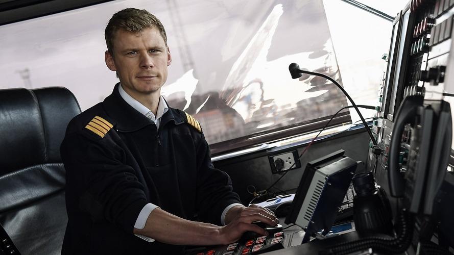 En mandlig kaptajn sidder ved kontrolpanelet på en skibbro, iført uniform med epauletter.