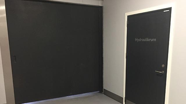 En sort skydedør og en hvid dør med teksten "Hydraulikrum" på en grå væg.