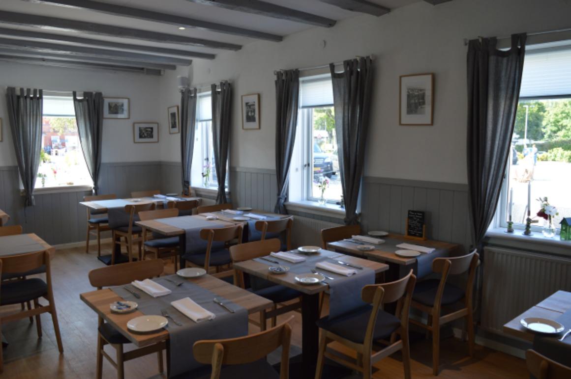 Indendørs restaurant med borde, stole og vinduer, dækket med grå gardiner og en lys atmosfære.