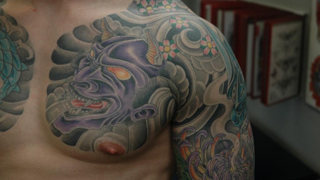 En mand med farverige tatoveringer på overkroppen, herunder en detaljeret drage og en ansigtstatovering.