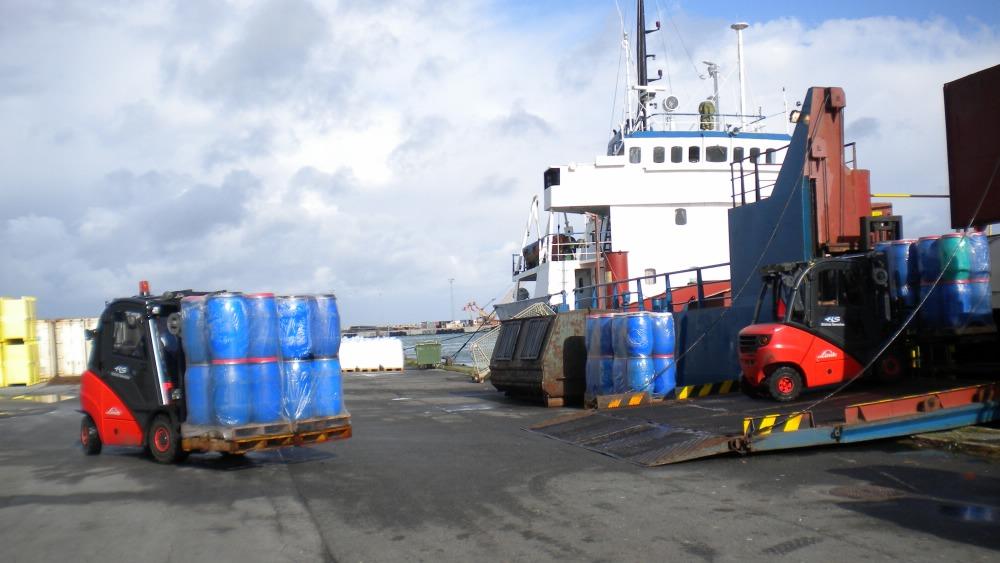 Lastvogne transporterer farvede tønder ved en havn med et skib i baggrunden.