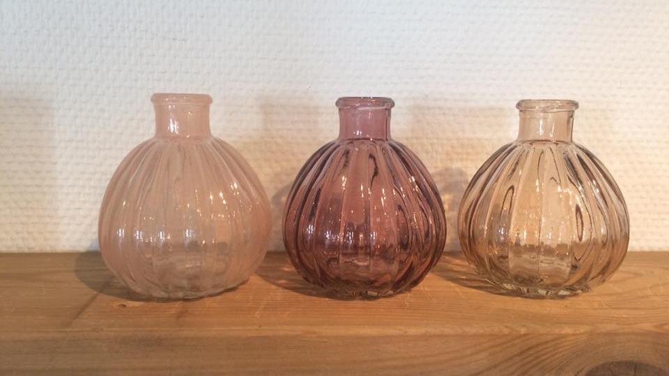 Tre farvede glasvaser i forskellige nuancer, placeret på en træhylde mod en hvid væg.