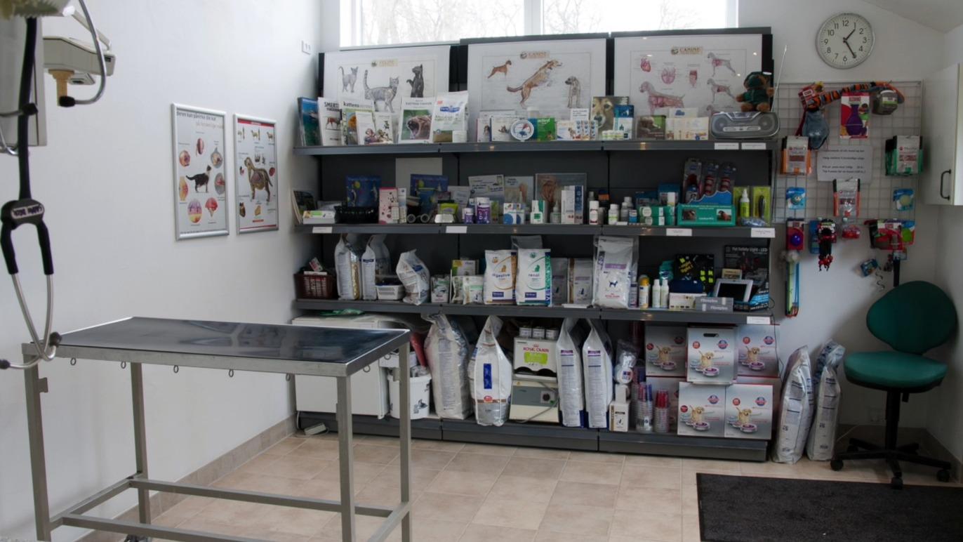 Dyrlægeklinik med udstyr, medicin og foder på hylder samt en behandlingsbænk i et lyst rum.
