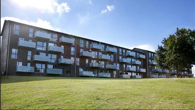 Moderne lejlighedsbygning med altaner, beliggende på en grøn græsplæne under en blå himmel.