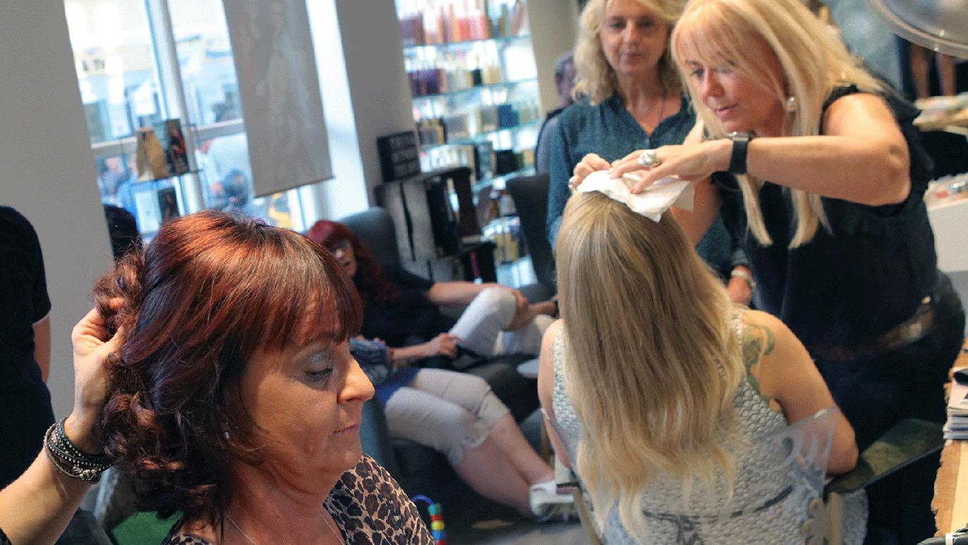 En frisørsalon med kvinder, hvor en frisør styler hår på en kunde, mens andre venter.
