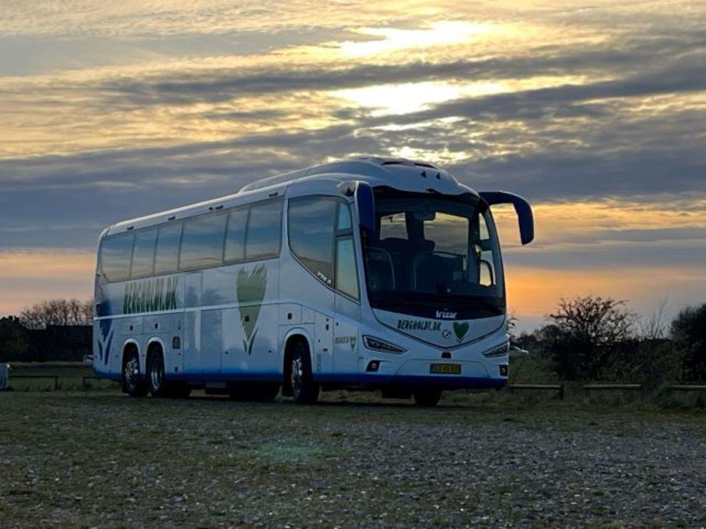 En moderne bus parkeret på en grusvej med en smuk solnedgang i baggrunden.