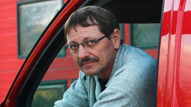 Mand med briller og mustache sidder i en rød bil, iført en grå sweater, med bygninger i baggrunden.