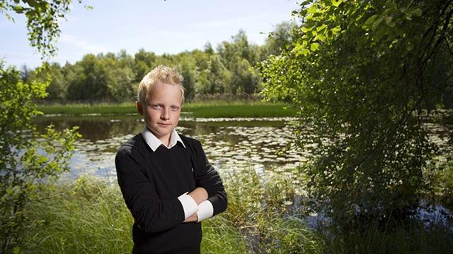 Dreng står med armene over kors ved en sø omgivet af grønne træer og græs.