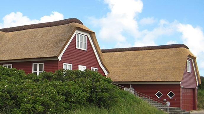 Rødt hus med stråtag og grønne buske i forgrunden under en blå himmel med skyer.