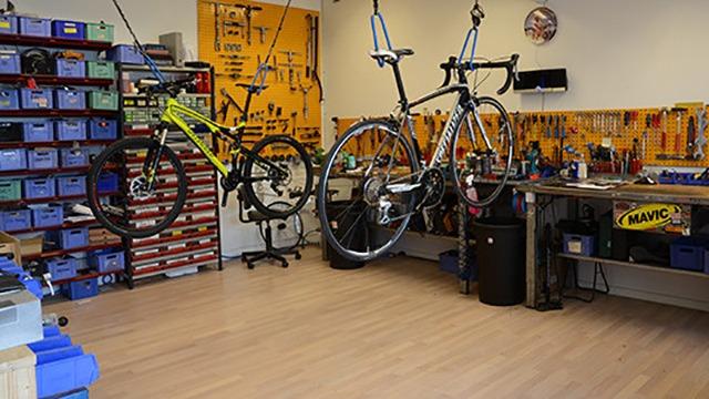 Cykelværksted med to cykler hængende, værktøjer og opbevaringskasser i baggrunden.