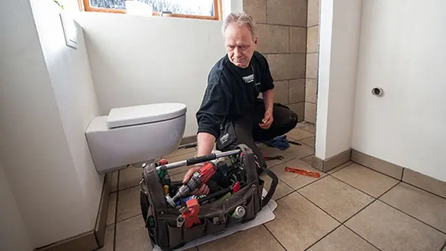 En håndværker arbejder i et badeværelse med værktøjskasse og toilet i baggrunden.