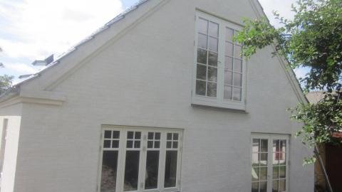 Hvidt hus med skråtag og store vinduer, omgivet af grønne træer og en blå himmel.
