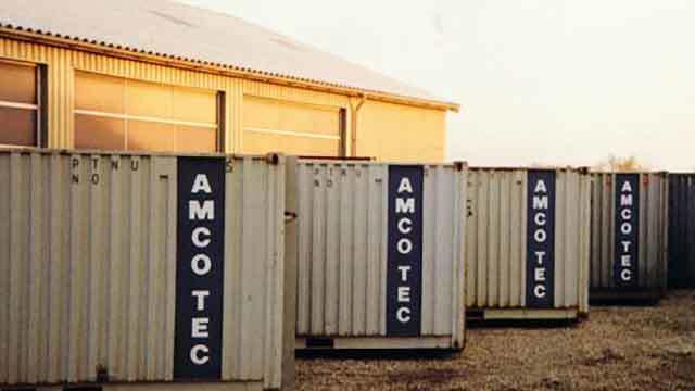 Lagercontainere med AMCO TEC-logo ved en bygning i solnedgang.