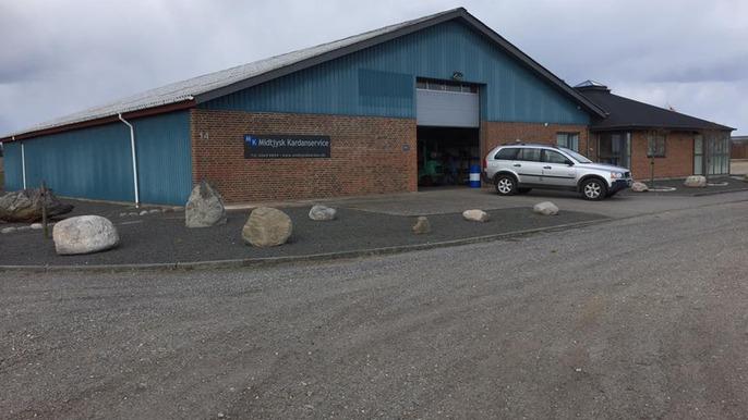 En blå bygning med murstensvæg og en grå bil parkeret foran, omgivet af store sten og grusvej.