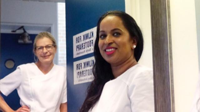 To kvinder i hvide uniformer står ved en dør i en klinik, smilende og klar til at hjælpe patienter.