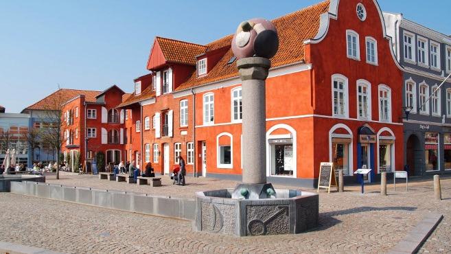 Farverig plads med røde bygninger og en skulptur i midten, omgivet af mennesker og bænke.