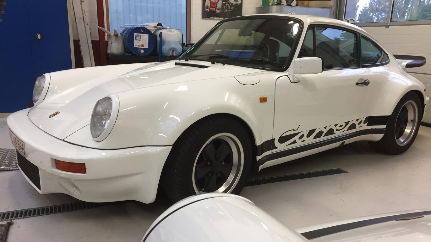 En hvid Porsche 911 Carrera med sort dekoration står i en garage med værktøj og udstyr i baggrunden.